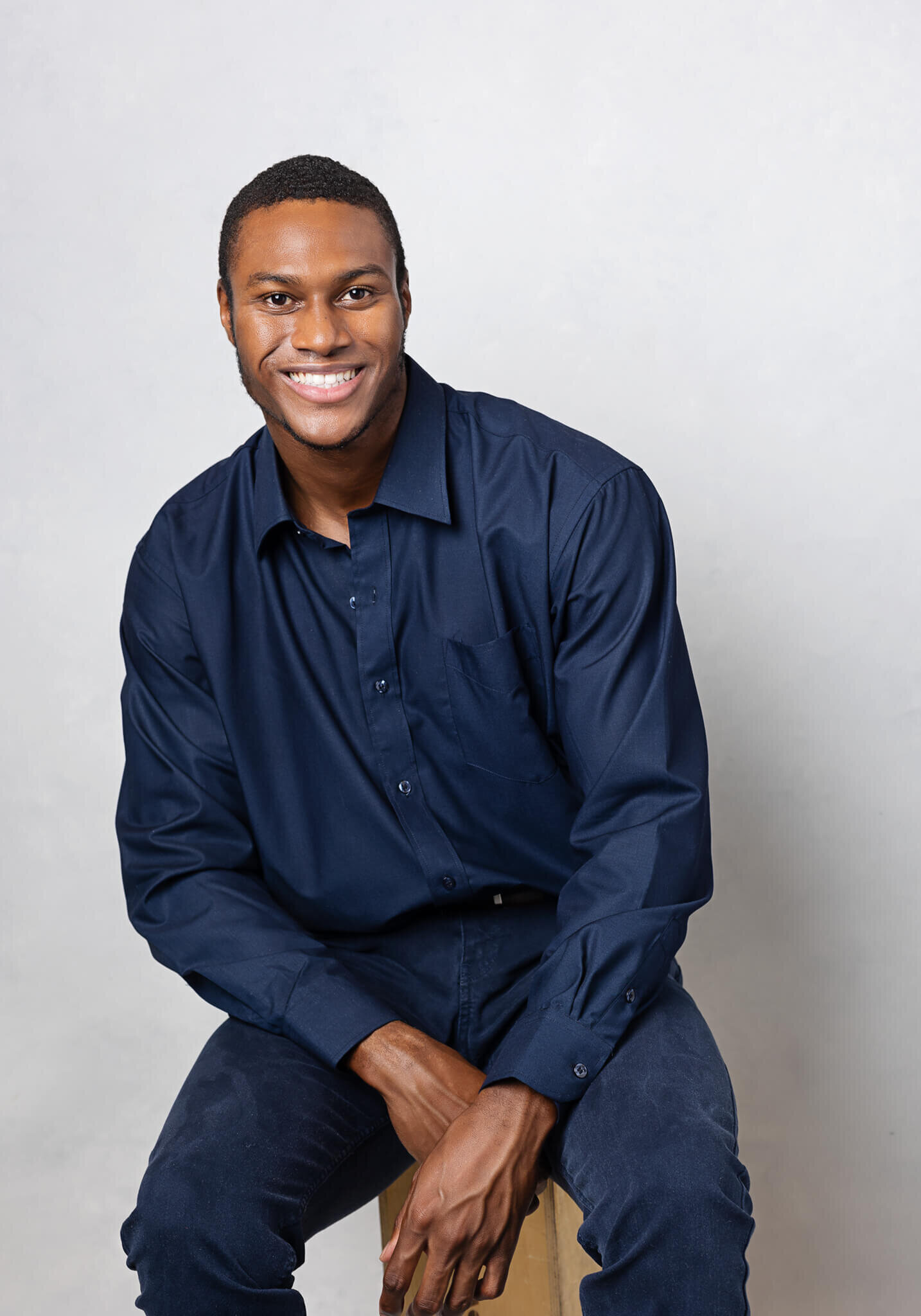 young professional is sitting on a chair and posing while looking relaxed and happy, he is wearing a blue shirt and jeans