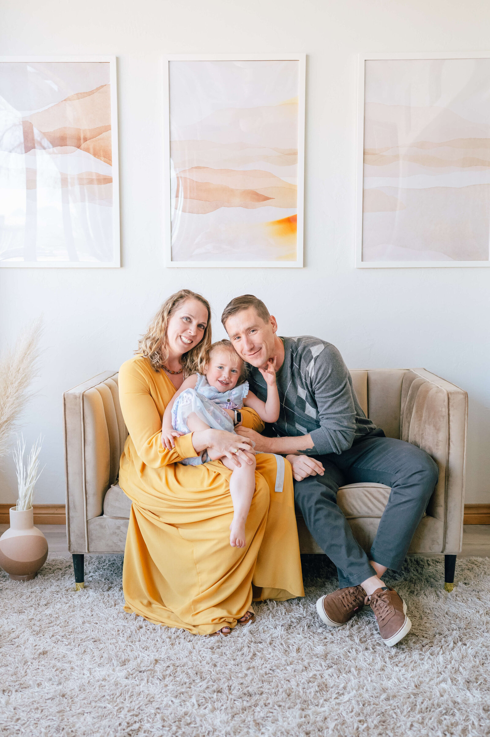 Within a white and well decorated room, a woman in a yellow dress holds her playful daughter while her husband hugs his daughter and looks at the camera