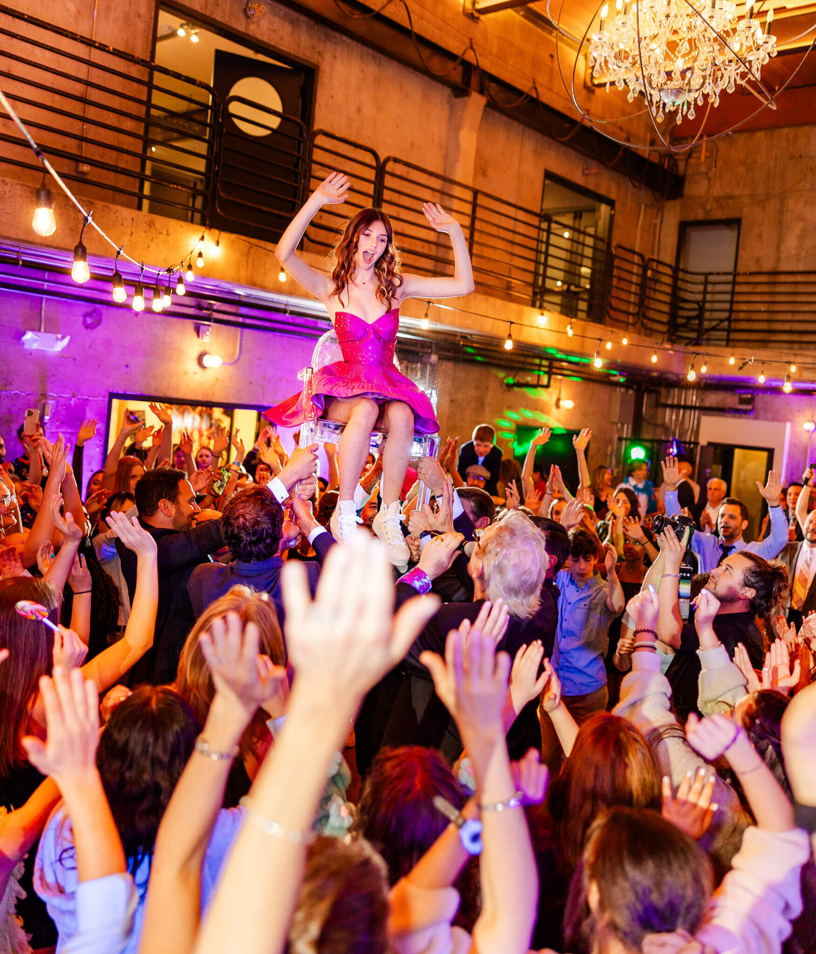 A teenage girl celebrates and dances while sitting on a lifted chair in the middle of a crowded dance floor