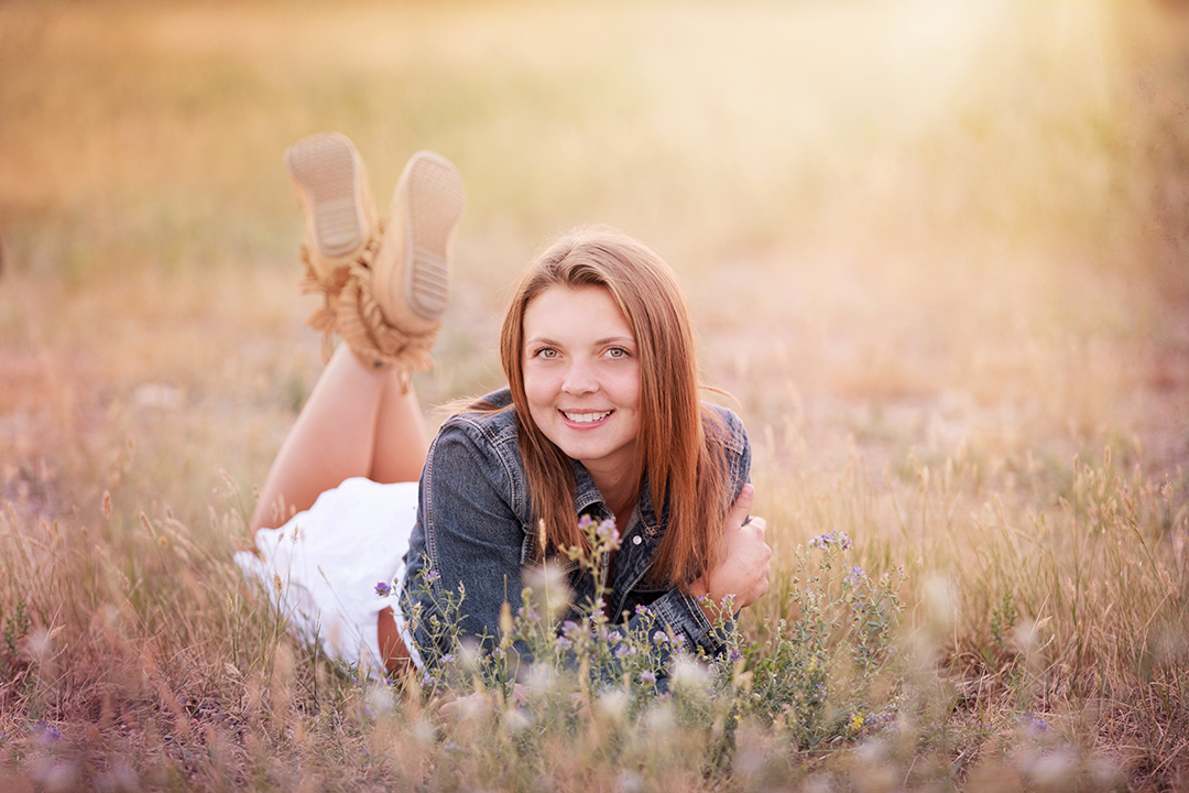 High School Senior pictures, Senior Pictures Near Me. graduation pictures, photographers in Billings, MT. Senior in sun kissed grassy field, wearing jean jacket and white skirt, brown hair smiling at camera.