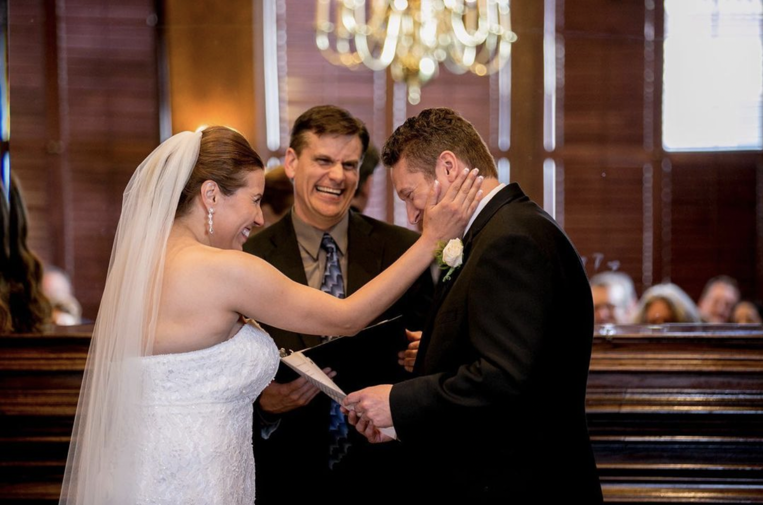 Bride laughs and holds groom's cheek while he reads his vows smiling