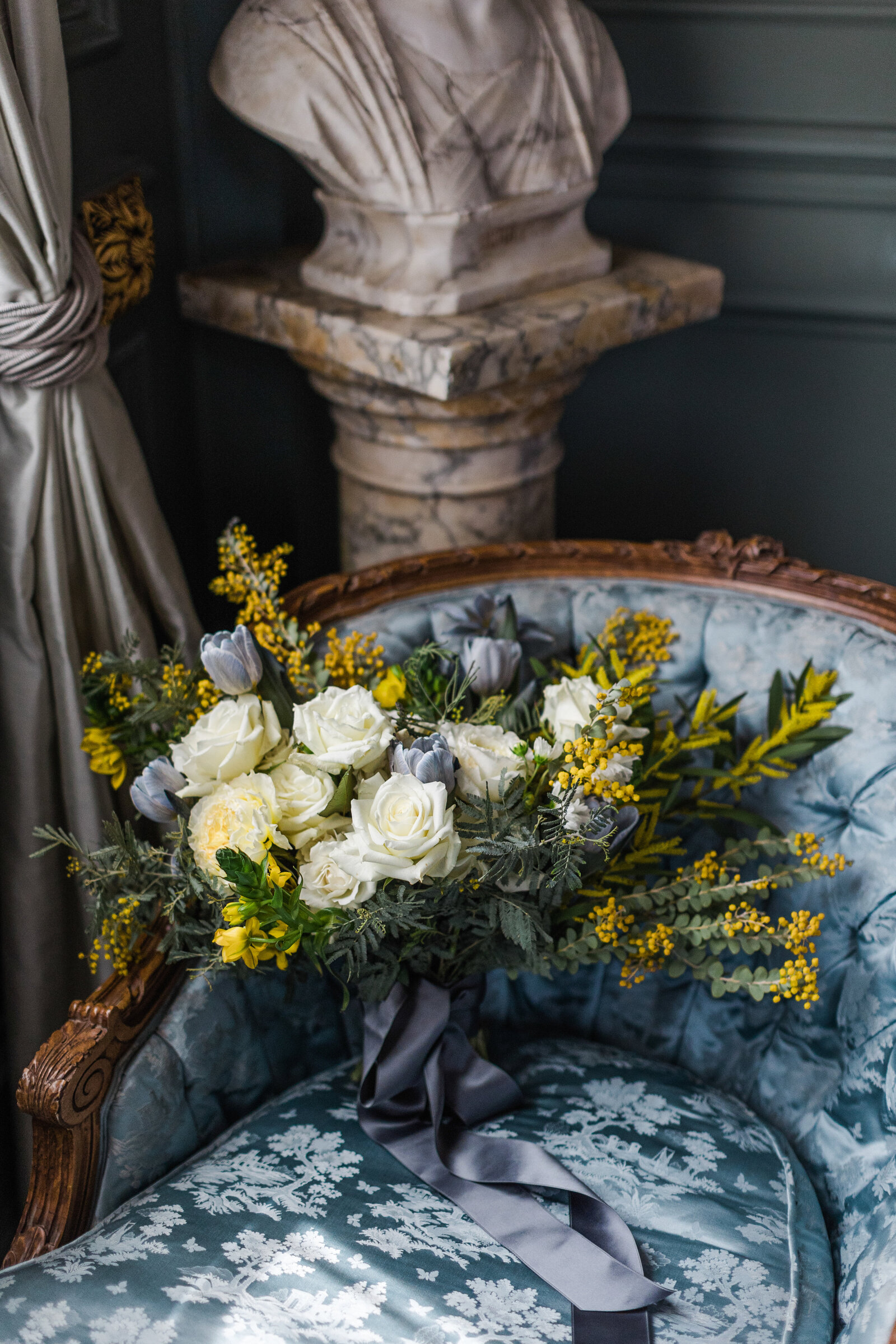 A detail shot of a wedding bouquet white and yellow flowers with blue ribbons flowing off the end. The bouquet is resting on a fancy blue chair with white floral patterns with a marble bust seen in the background.
