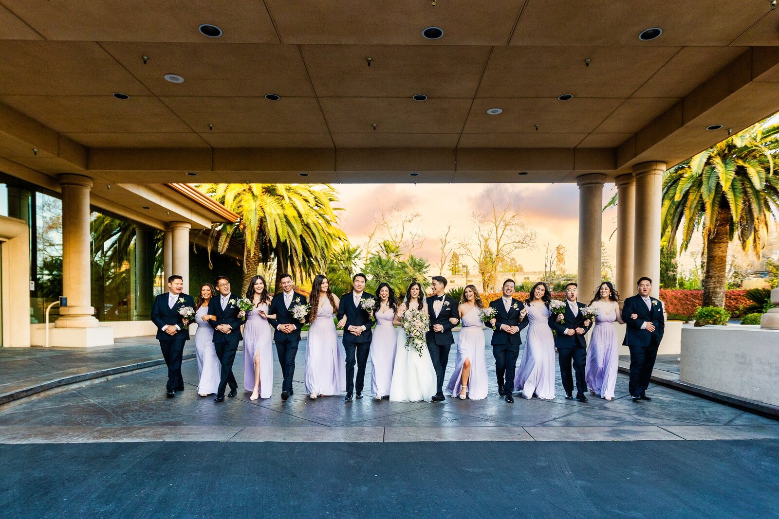 Bridal wedding party walks with each other arm in arm at Arden Hills in Sacramento under their awning with palm trees in the background. Photo by Sacramento wedding photographer, Philippe Studio Pro.