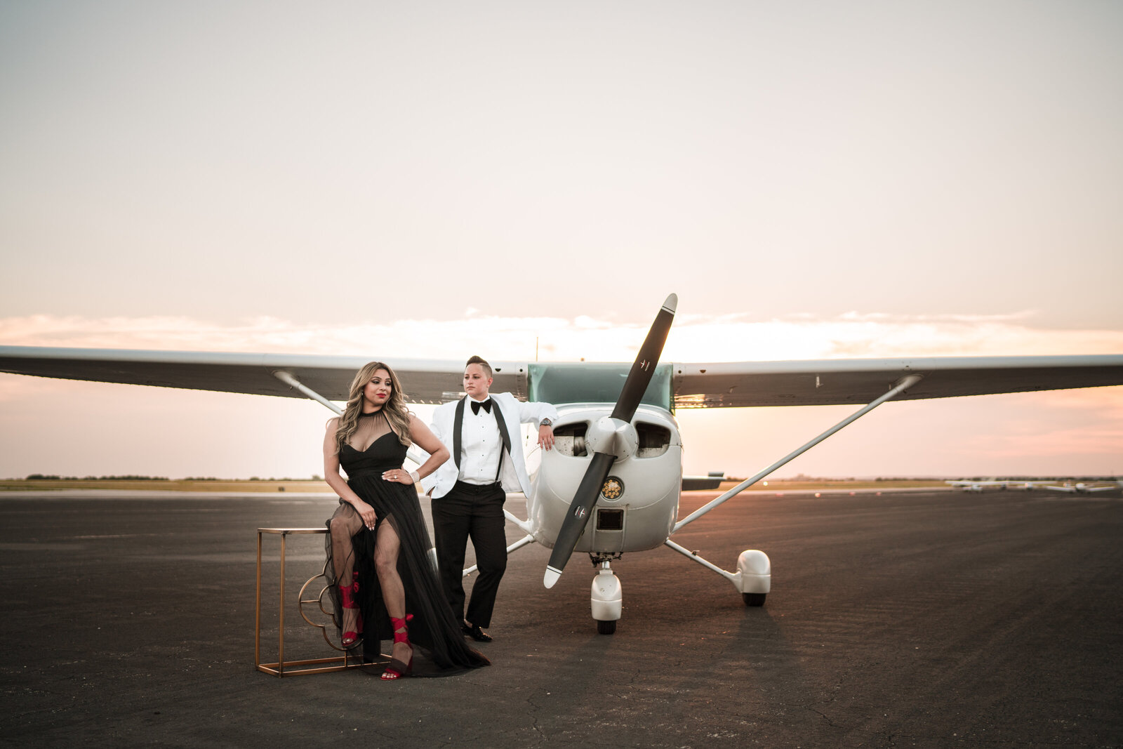 Gay couple's engagement session wearing a tuxedo and black gown standing in front of an airplane, in San Antonio.