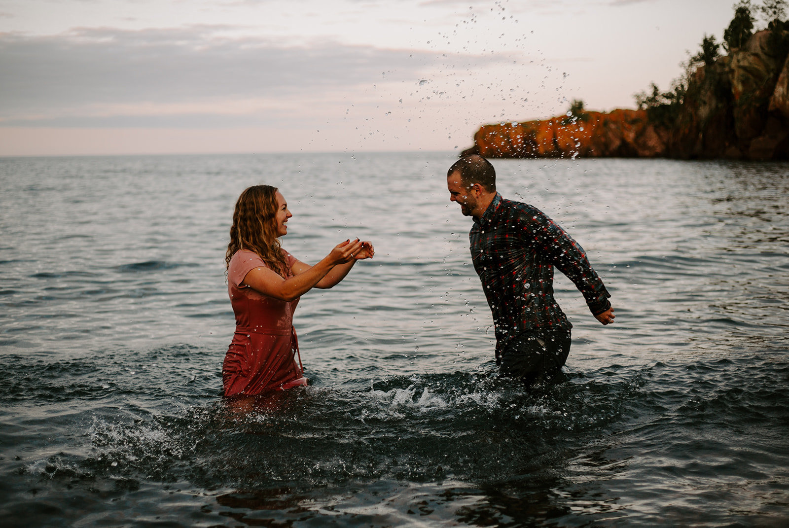 Couple splashing each other playing in the water at the North Shore in Minnesota during their engagement session