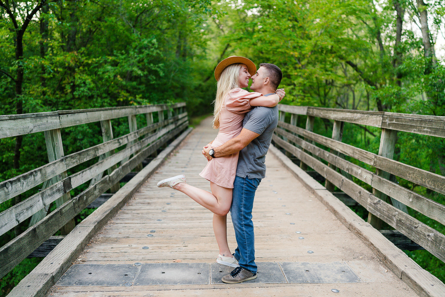 Man lifts woman dressed in pink dress and hat on a bridge.