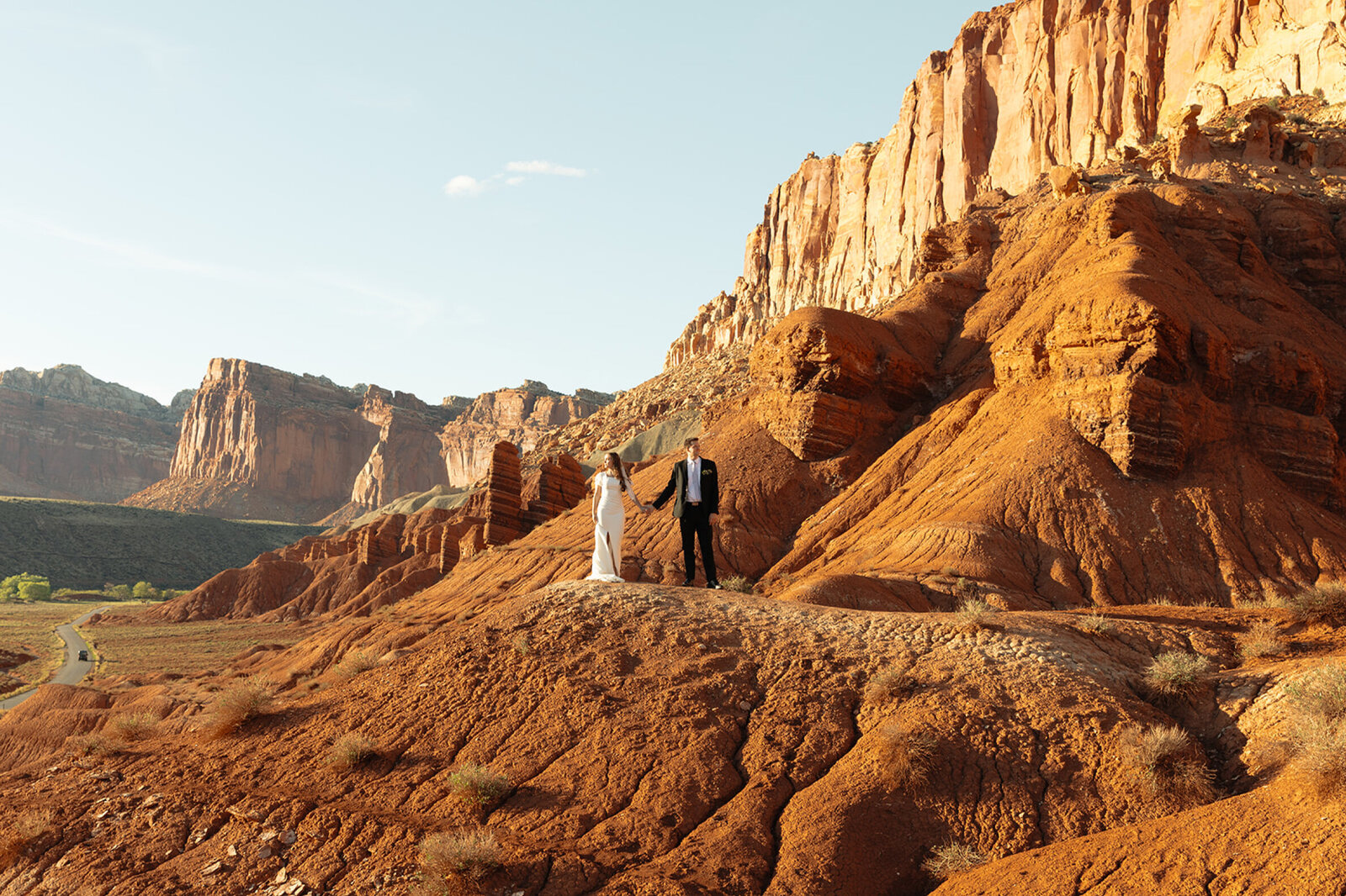 A distant shot of a bride and groom holding hands and looking in opposite directions to the left and the right. They are standing on beautiful red rocks with Capitol Reef's magnificent buttes and stunning landscape behind them.