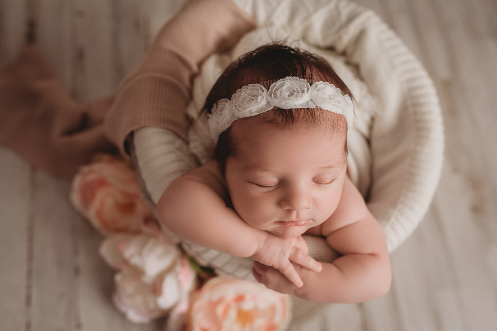 Newborn girl portrait in basket on white flooring with chin on hands and flower headband. Photo shot from above and looking down at baby girl.