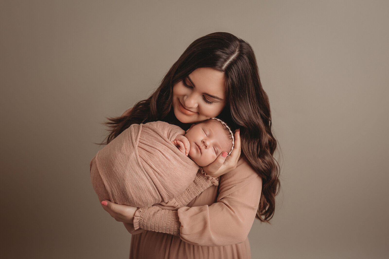 Long brown haired mom in mauve long sleeve gown holding and snuggling newborn baby girl wrapped in pink