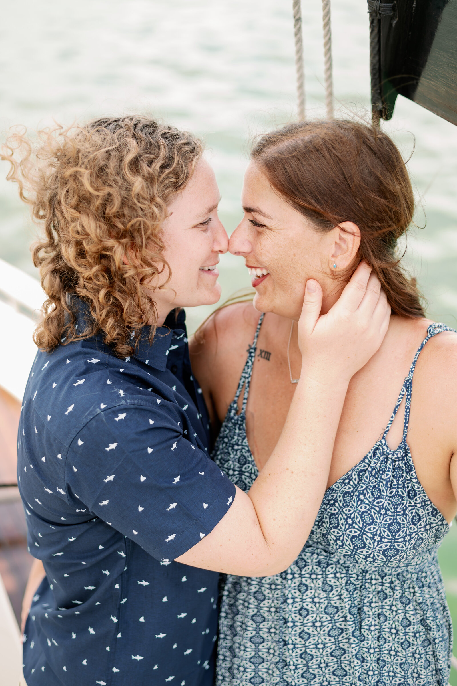 engaged couple smiling and touching noses, one with a hand placed delicately upon the others neck to pull her in close with water behind them.