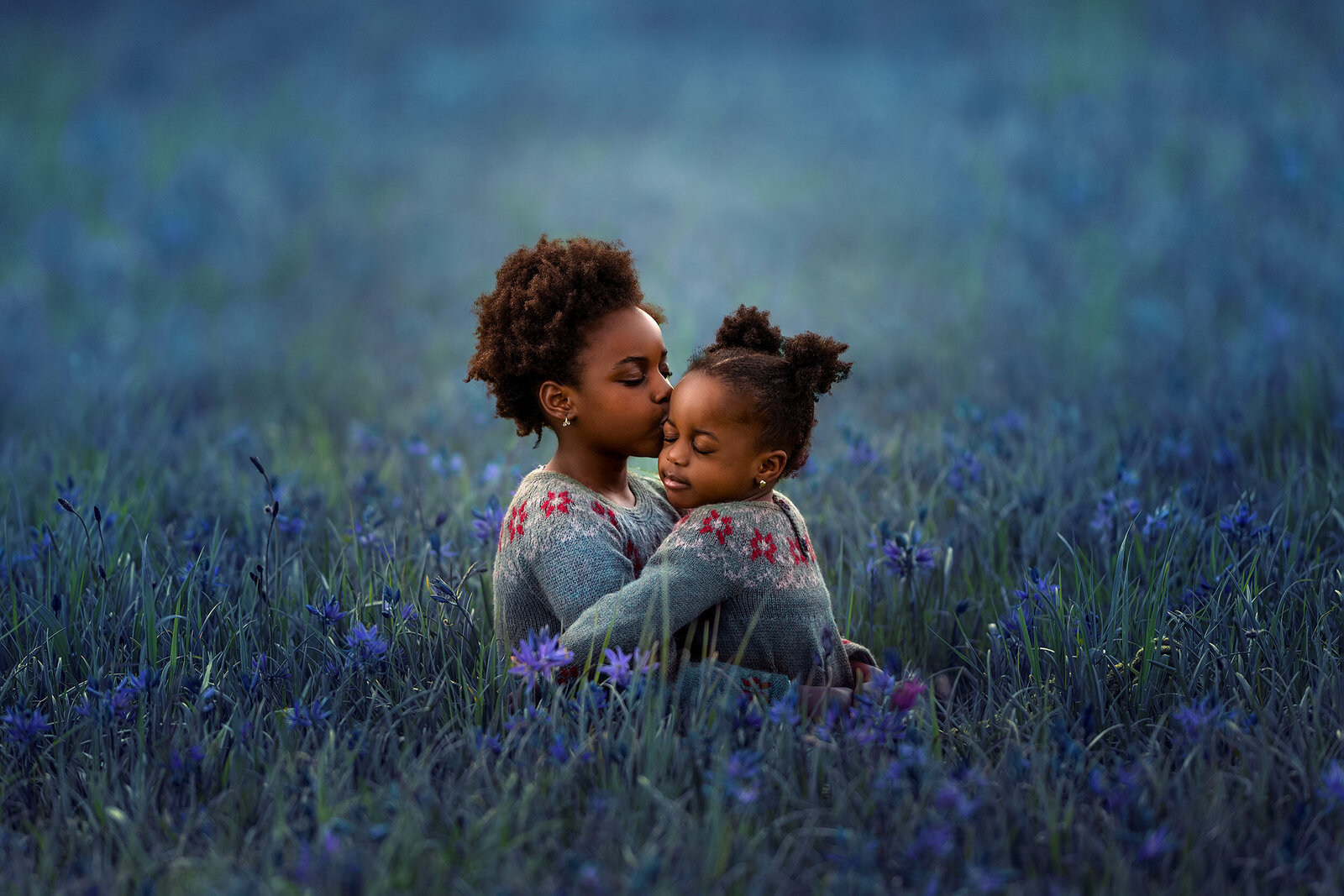 Two Black sisters share an embrace in a field of blue wildflowers.
