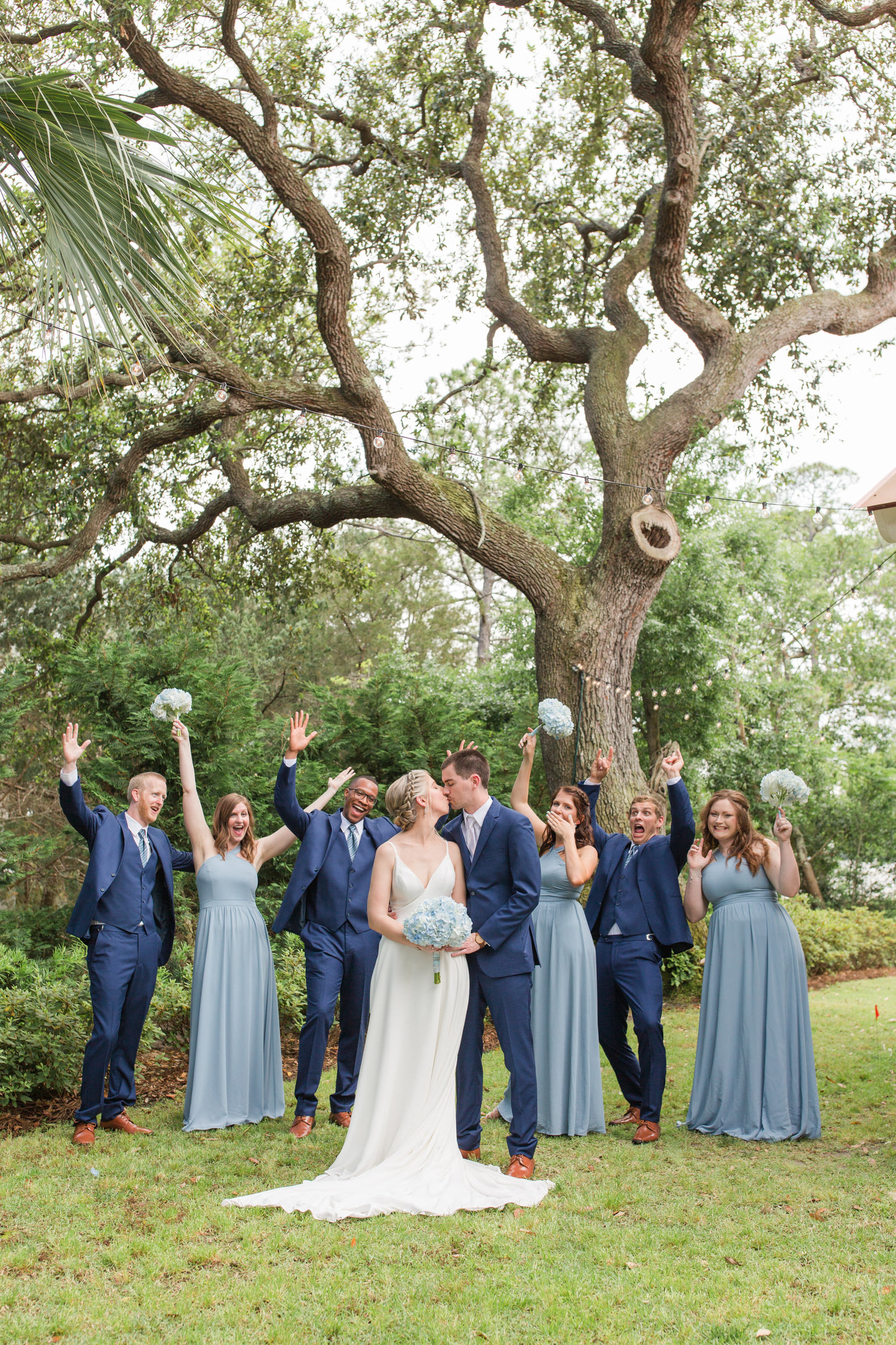 wedding party in blue dresses in front of oak trees cheering as bride and groom kiss