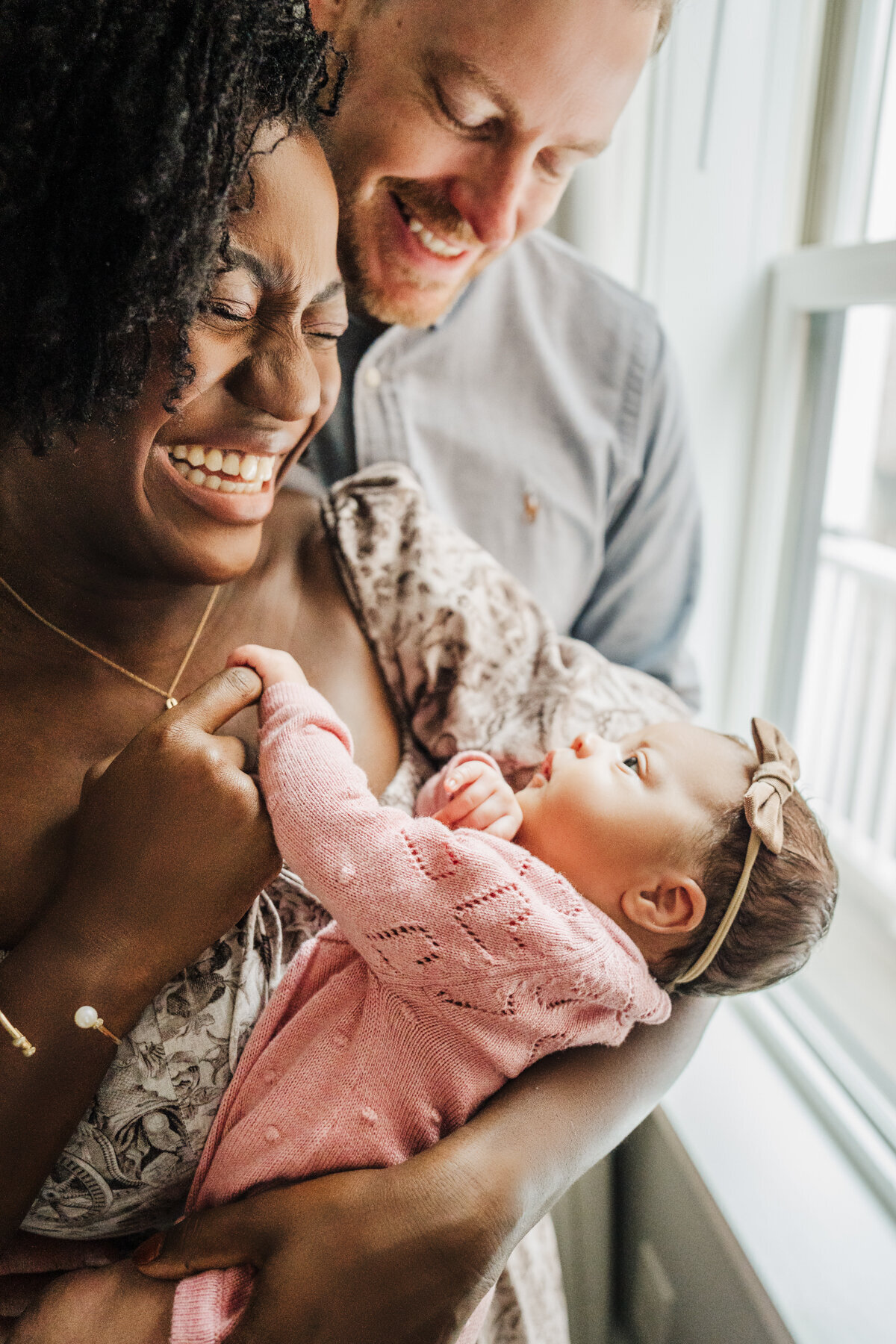 mom and dad stand by window and laugh together while holding infant girl