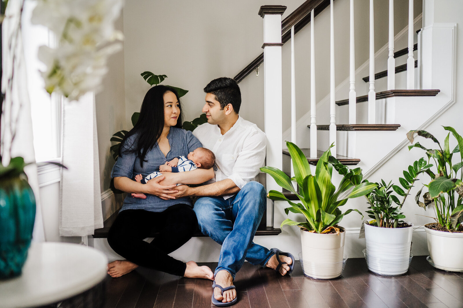 parents with new baby sit at bottom of stairwell surrounded by plants