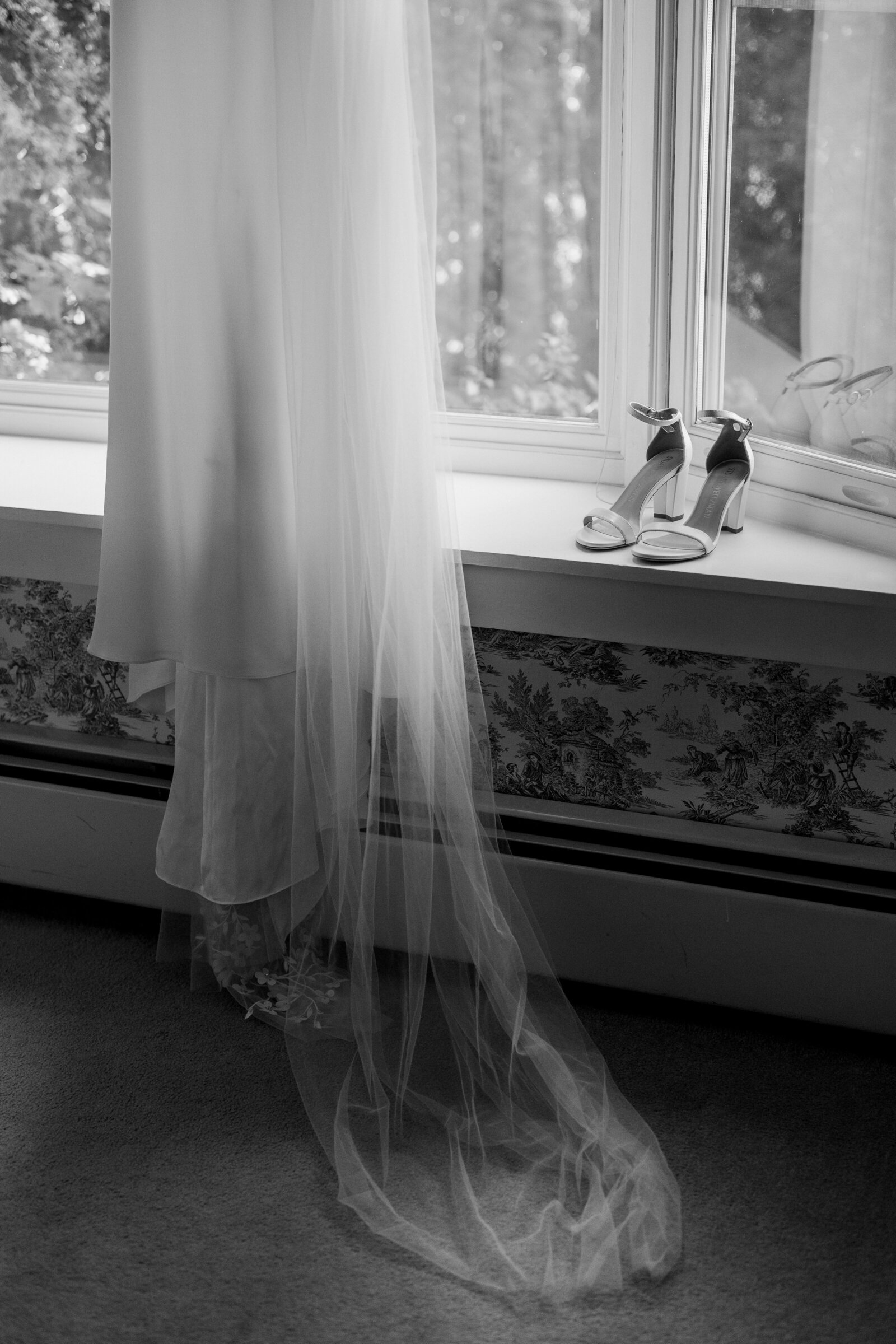 Black and white photo of heels sitting on a nook by a window with a wedding dress hanging next to them.