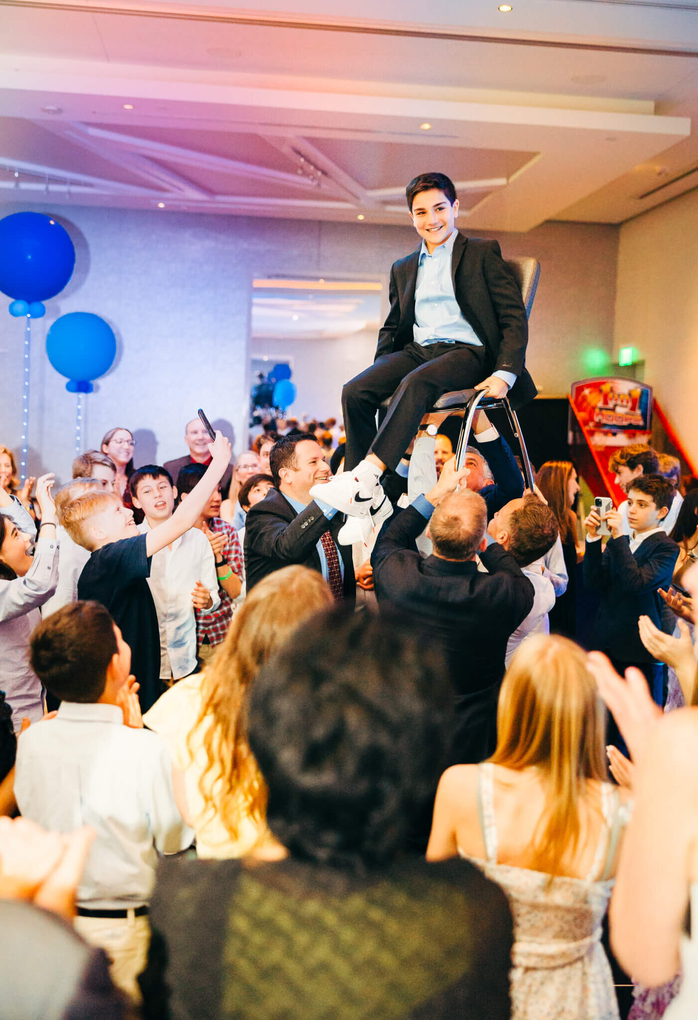 A teen boy in a black suit sits on a lifted chair in a crowded dance floor during some Bellevue Bar and Bat Mitzvah Photography