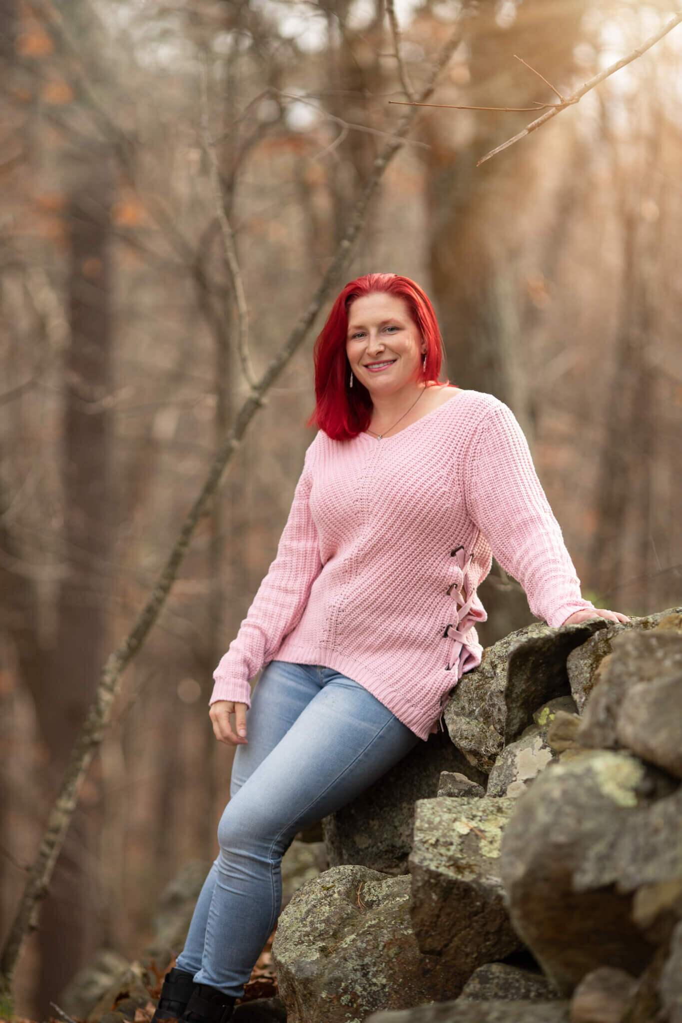 Woman ditting on rock wall in woods in New Hampshire with bright red hair and wearing a pink sweater