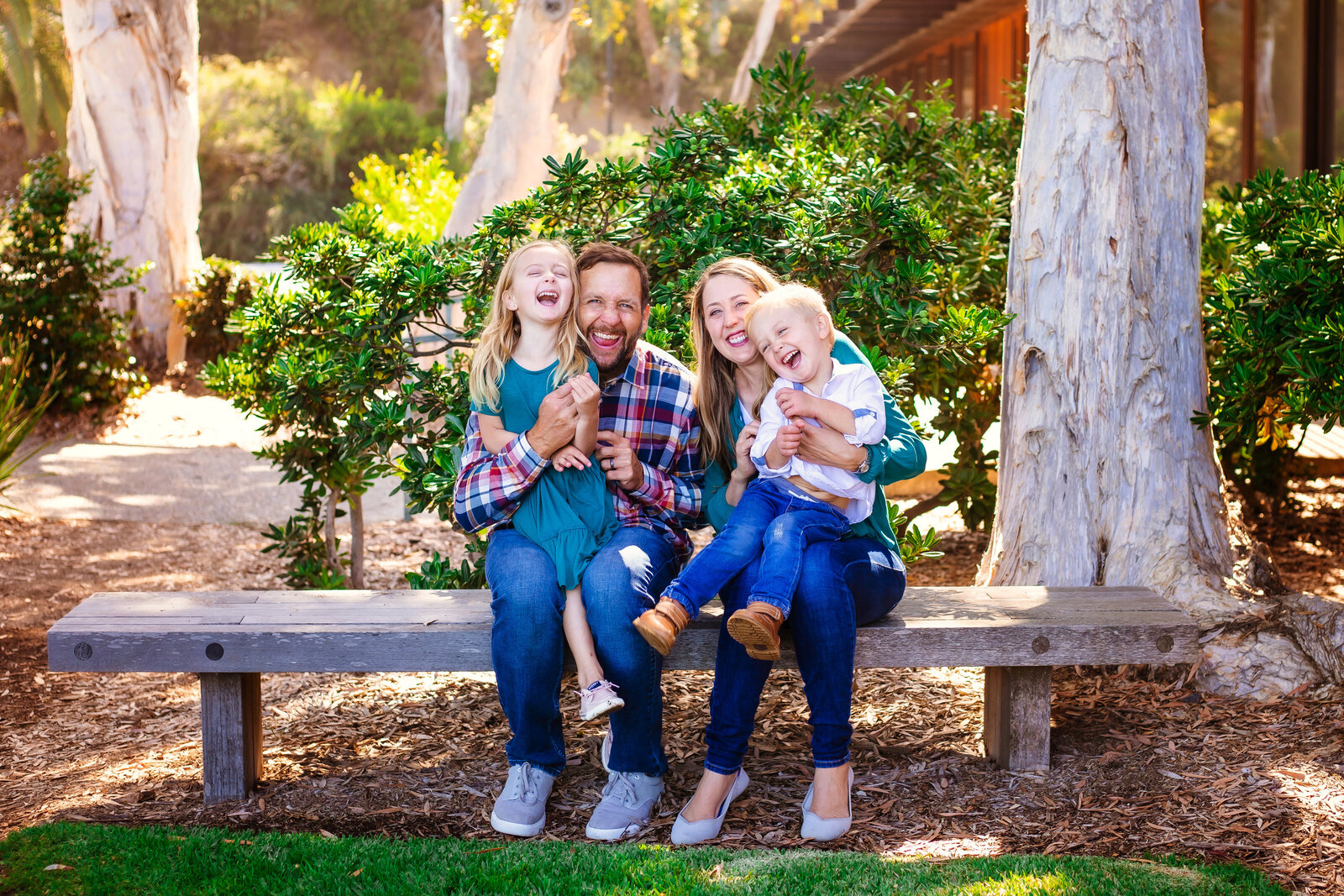 Family Photographer, a family of four with two young giggling daughters sit on a park bench