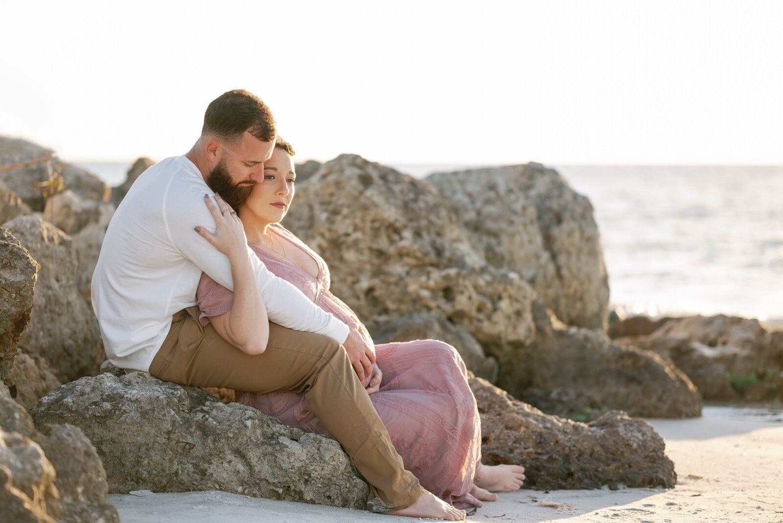 Pregnant woman snuggling with her husband on a rock jetty near the waterline of a beach