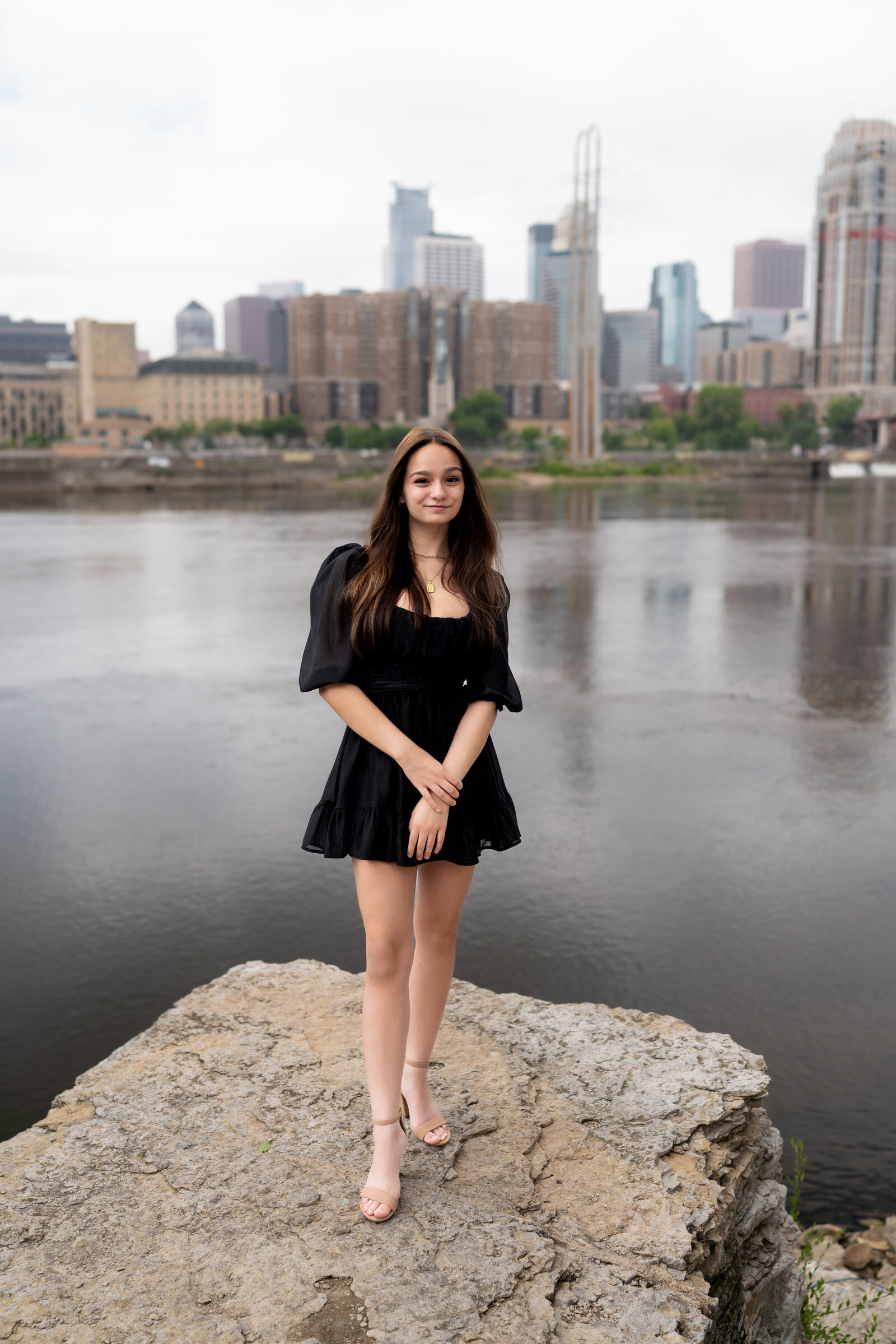 Senior girl in black dress stands on rock and smiles with Minneapolis in the background.