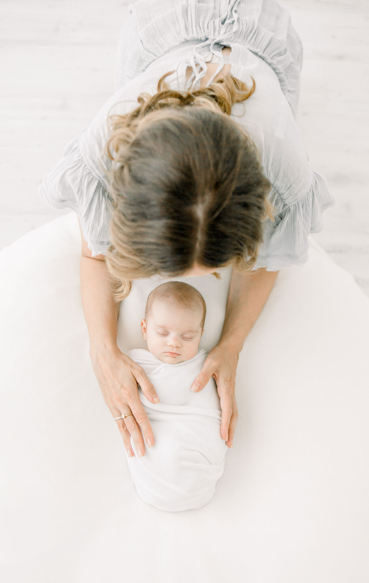 Neugeborenenfotografie aus der Vogelperspektive. Schlafendes Neugeborenes in weißer Decke. Die Mutter über dem Baby gebeugt und Hände aufgelegt.