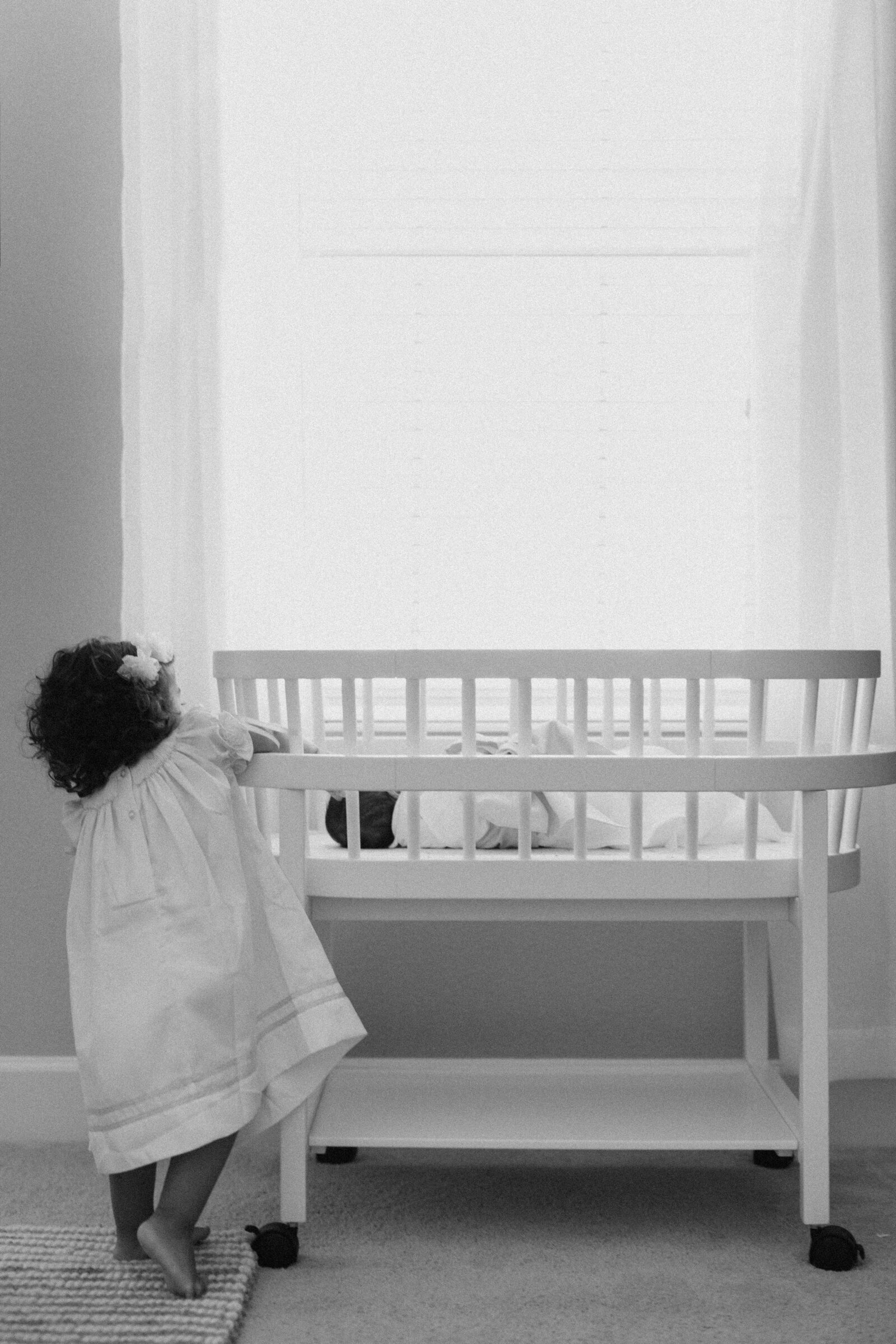 A new big sister stands on tip toes trying to reach into her baby brother's crib