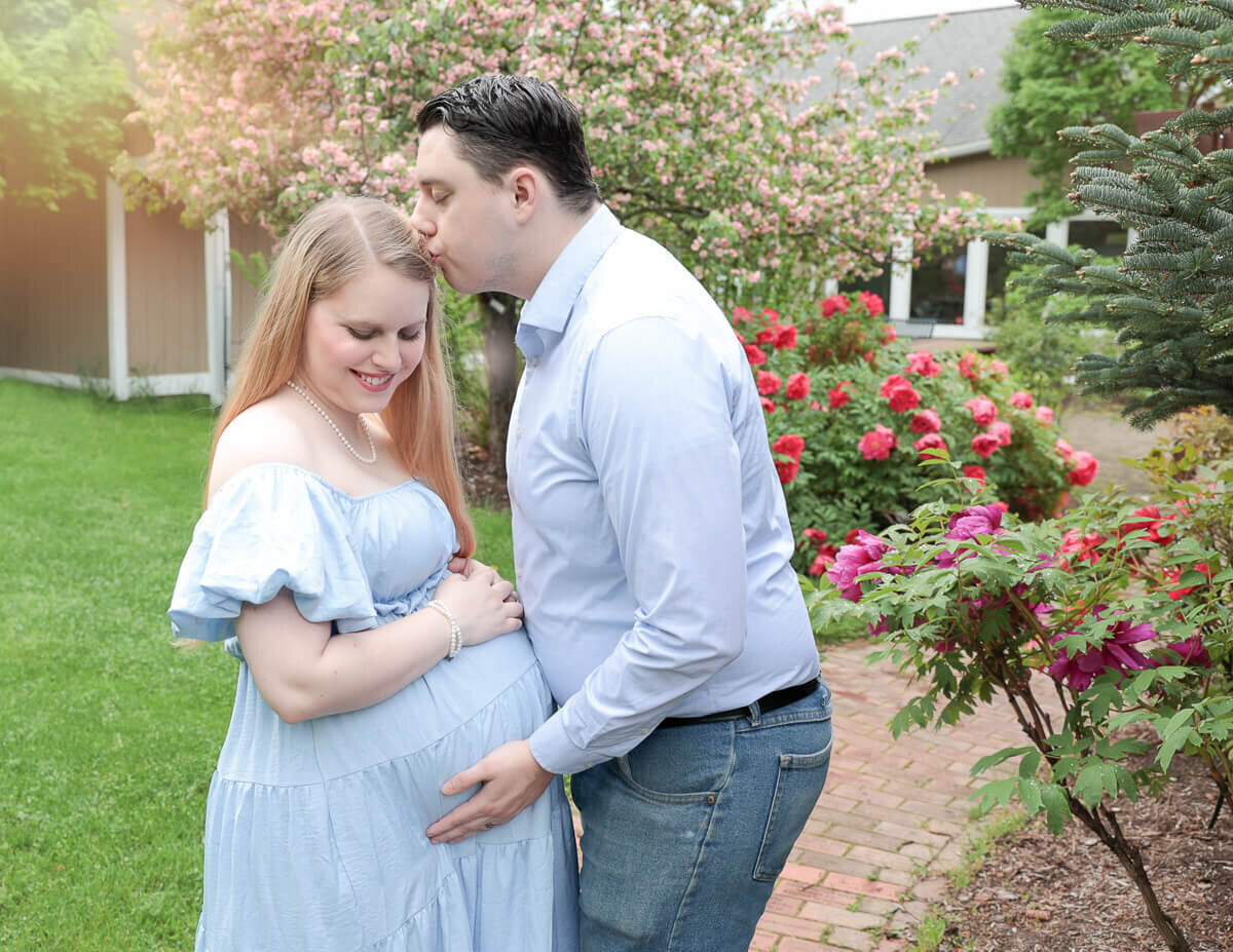 A father gently kisses the forehead of his wife while tenderly holding her pregnant belly in an outdoor setting, showcasing a beautiful maternity moment in Rochester, NY.