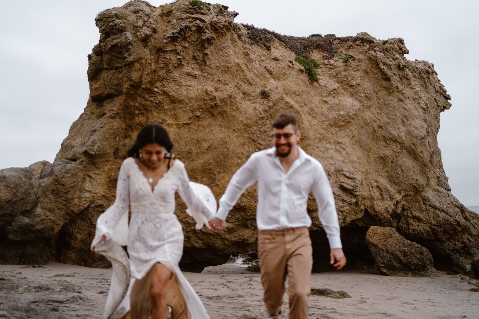 Couple holding hands and running on the beach in front of a large rock formation, bride in a lace dress and groom in a white shirt and tan pants.