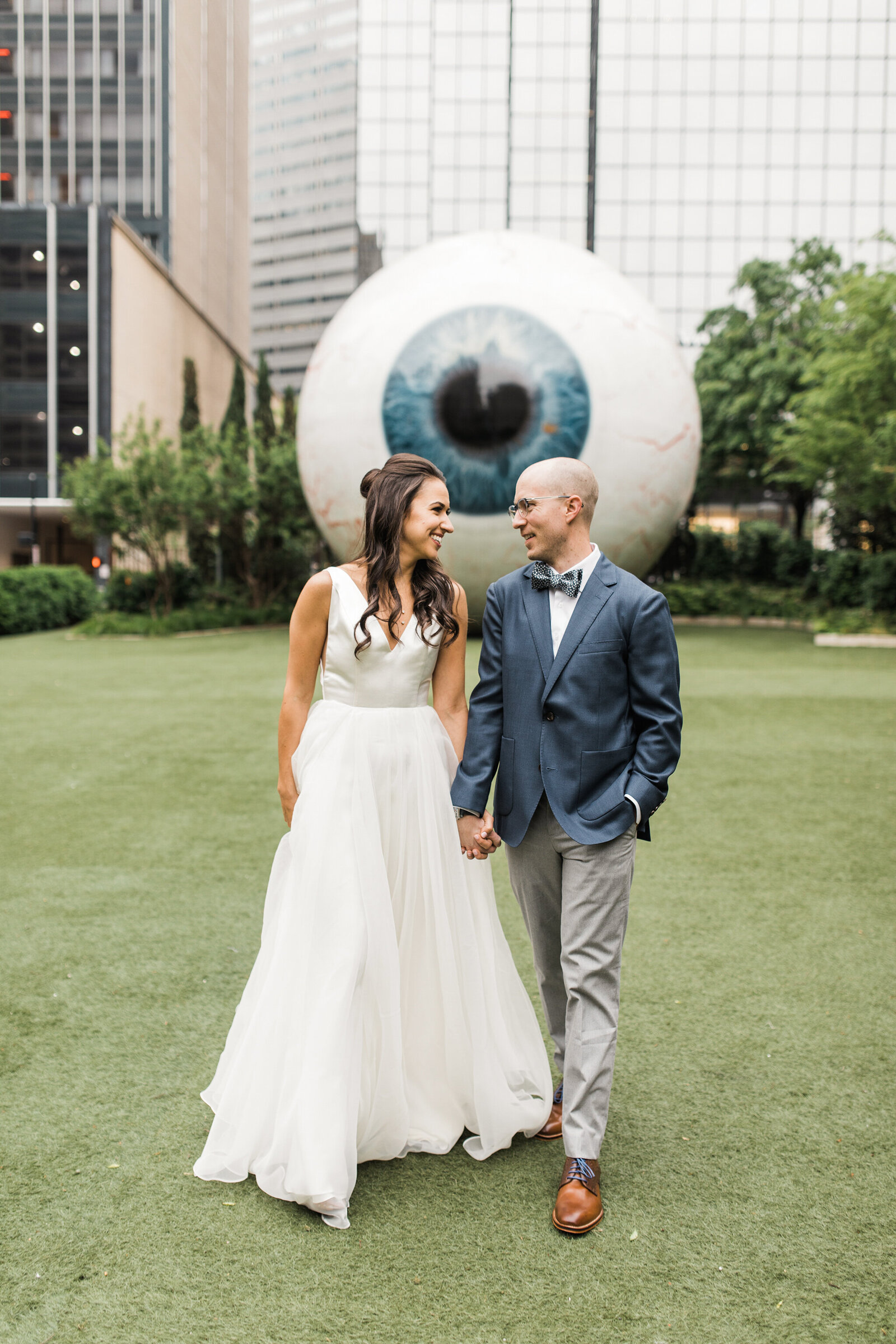 A couple standing in front of the Giant Eyeball in Dallas, Texas across the street from the Joule hotel in Dallas, Texas. The bride is on the left is wearing a sleeveless, long, white dress. The groom is on the right and is wearing a blue suit with a bowtie.