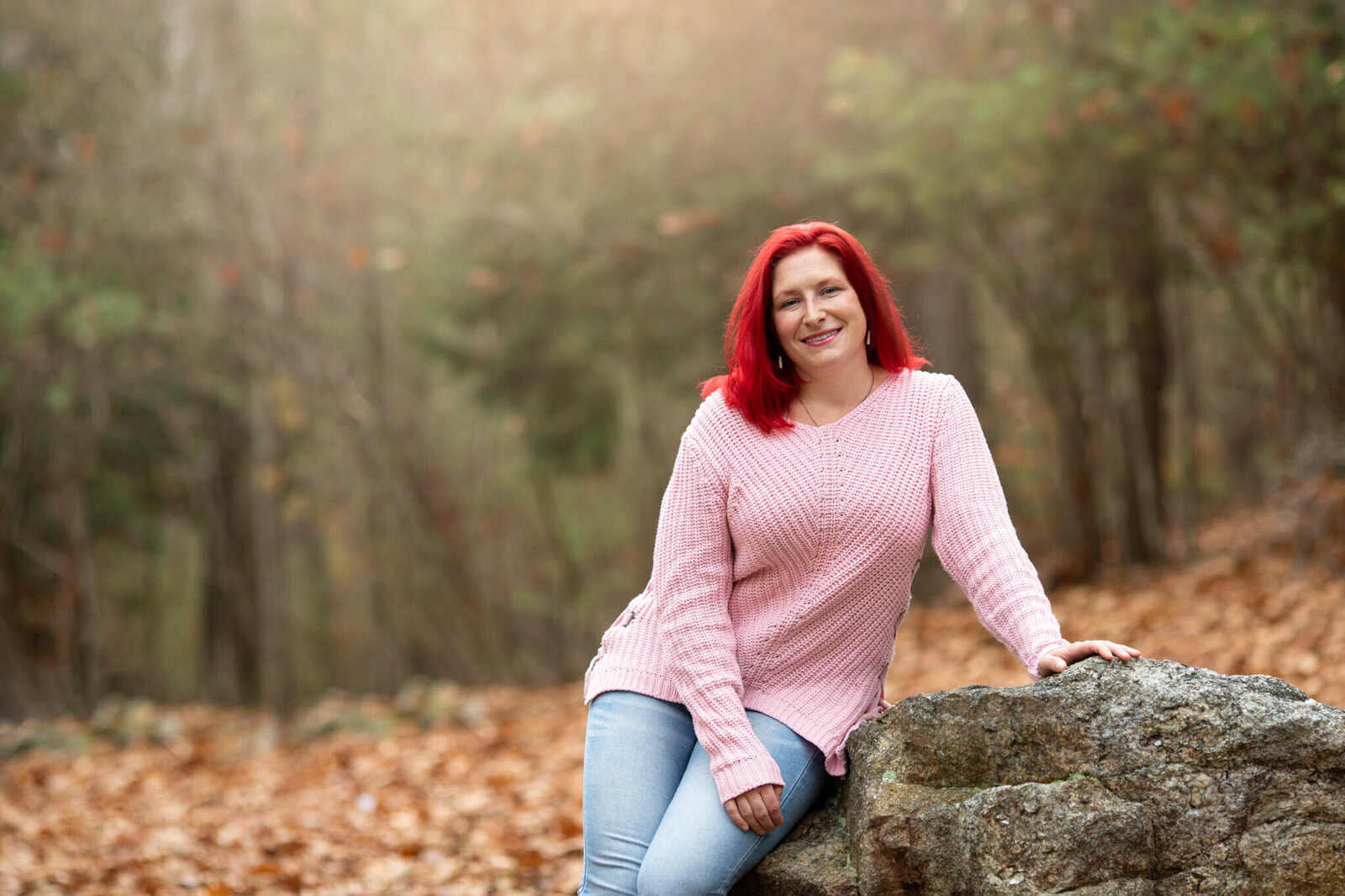 Woman ditting on rock wall in woods in New Hampshire with bright red hair and wearing a pink sweater facing camera