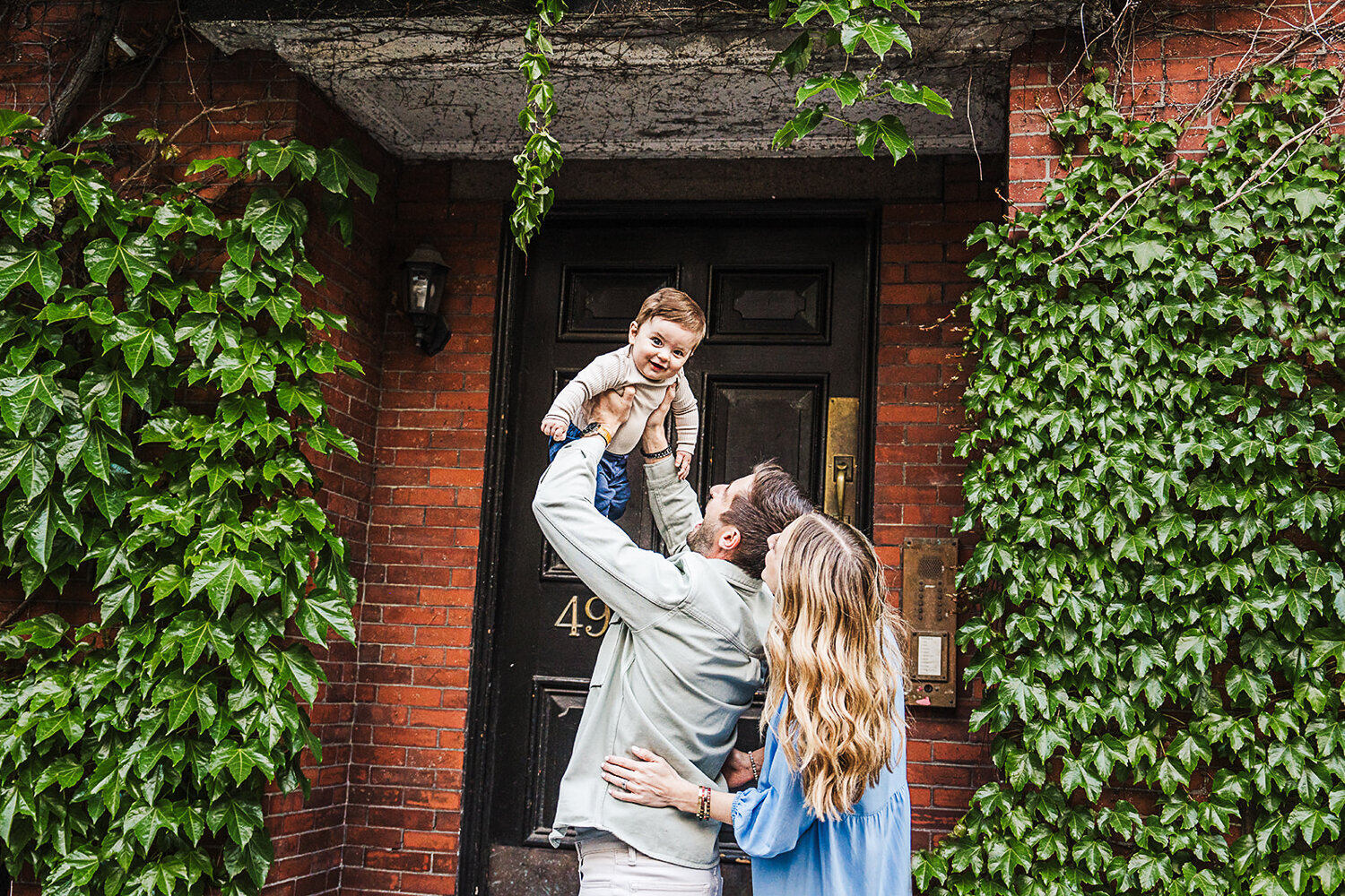 couple lift baby in air next to a boston wall of ivy