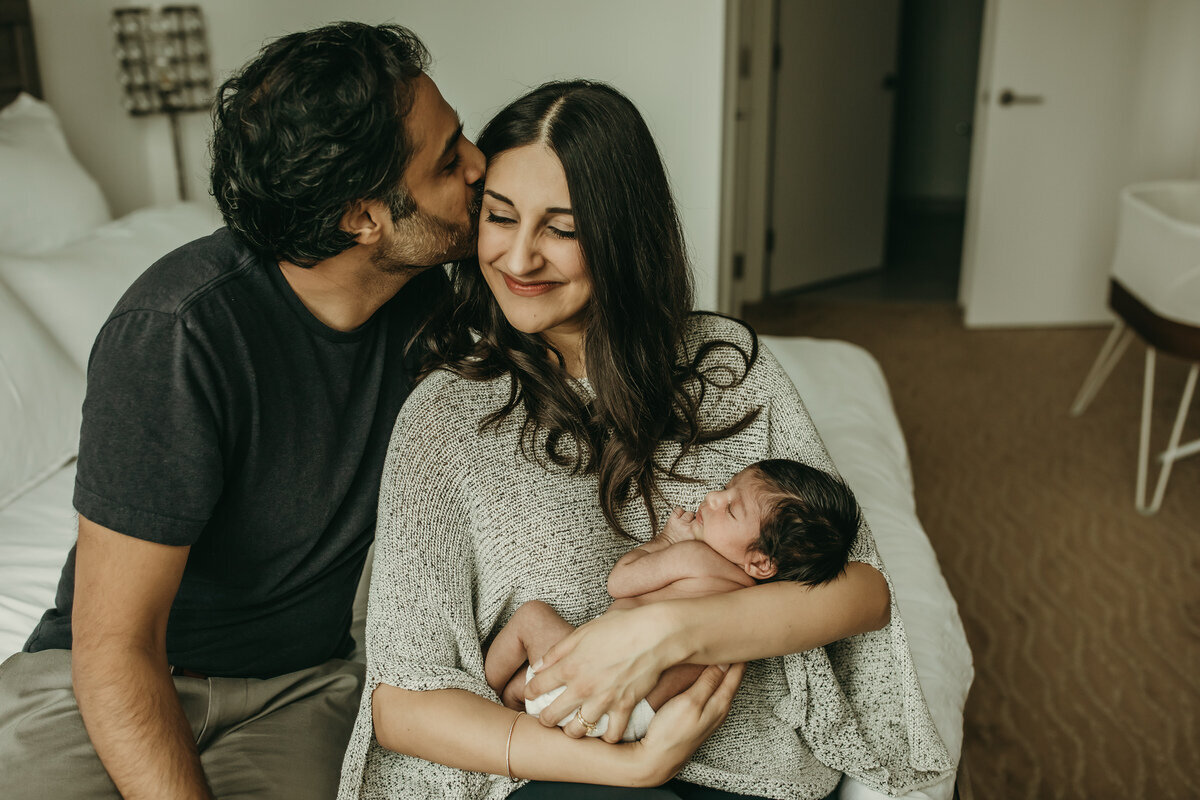 In home lifestyle newborn session with dad kissing moms cheek while she holds baby