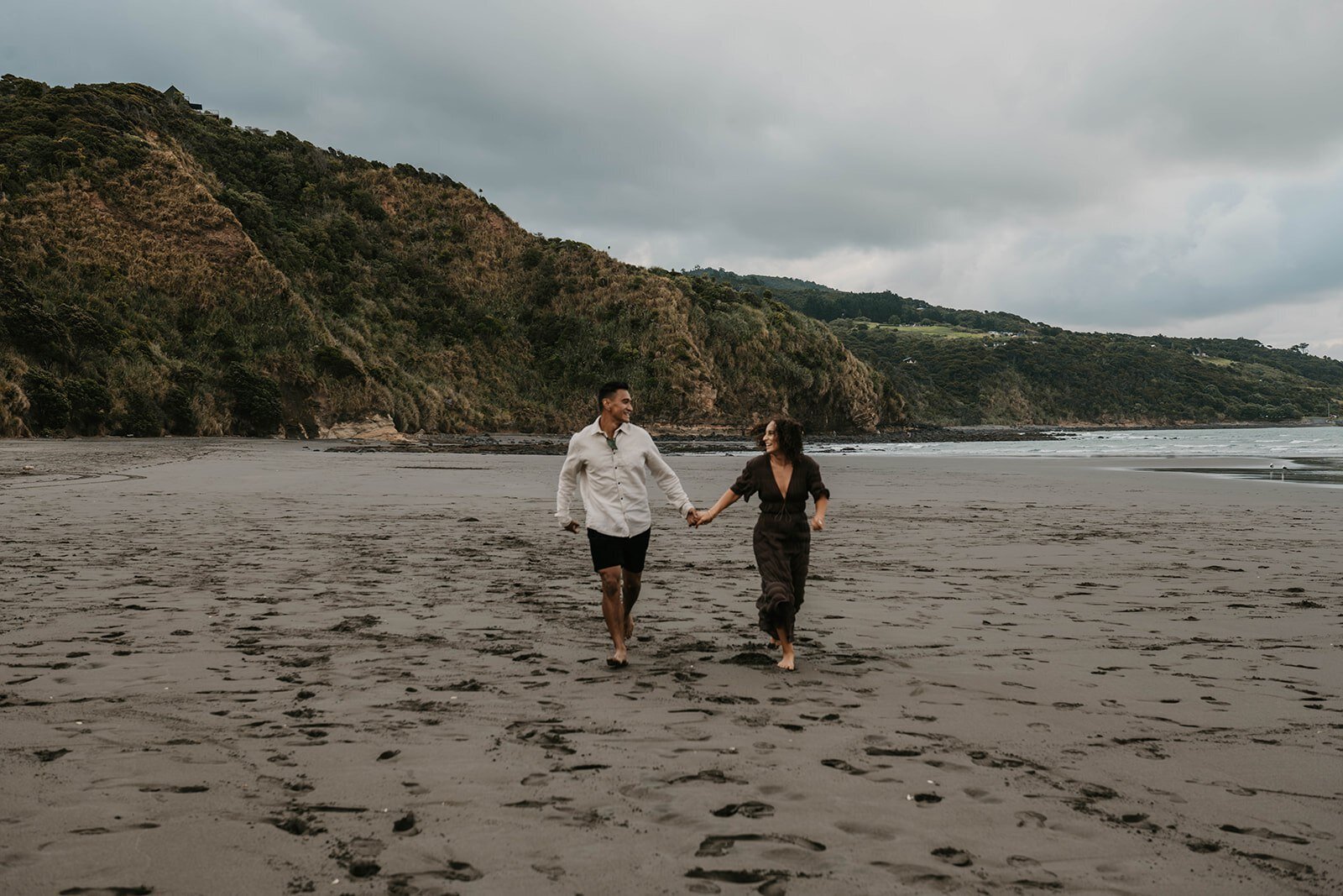 A couple holding hands and walking on a sandy beach with green hills in the background, evoking the spirit of an Italy photography retreat in the countryside for inspired storytelling.