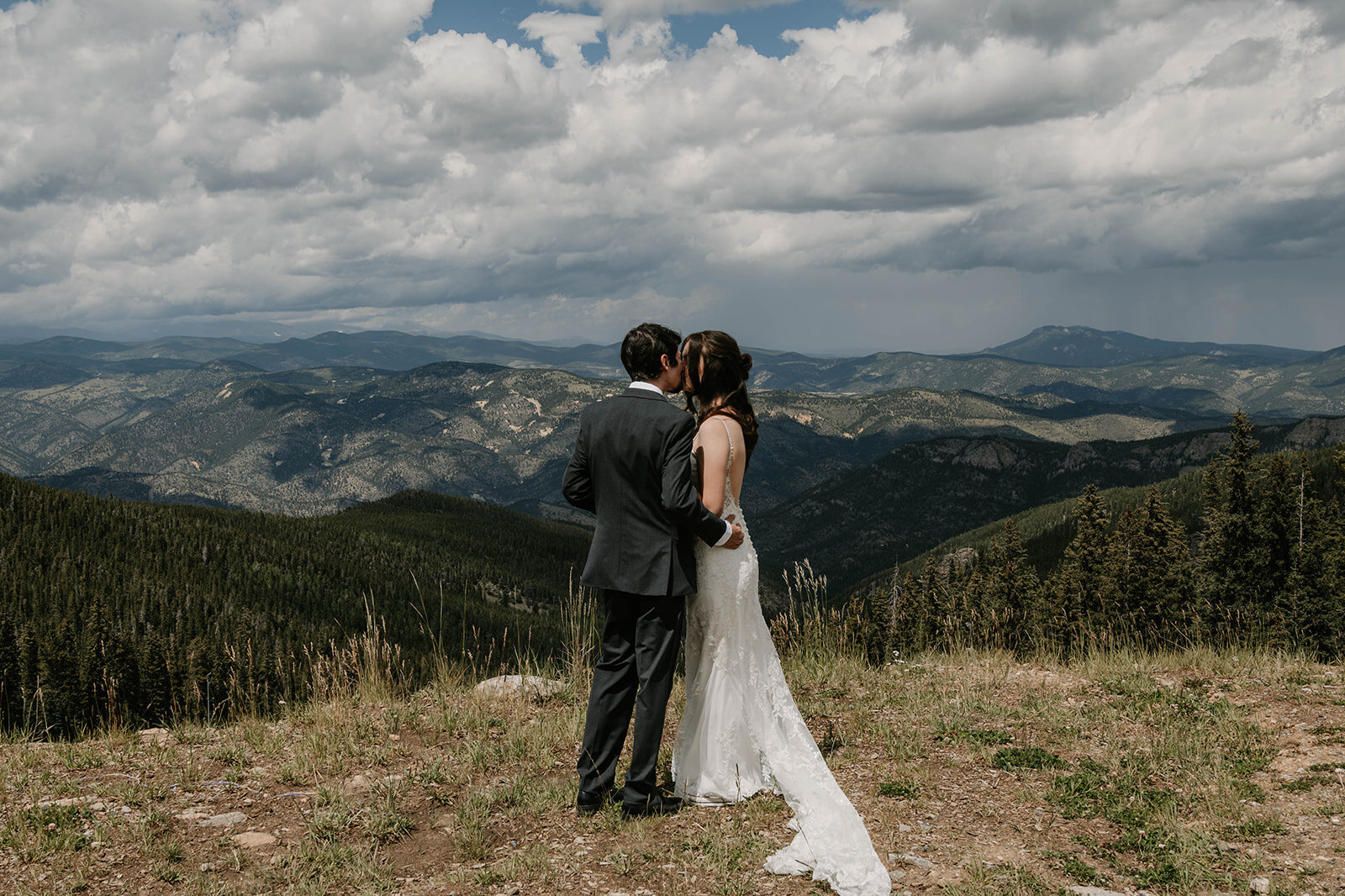 A bride and groom kiss on a mountaintop beneath dramatic clouds, forested hills behind them—captured in candid wedding photography at golden hour.