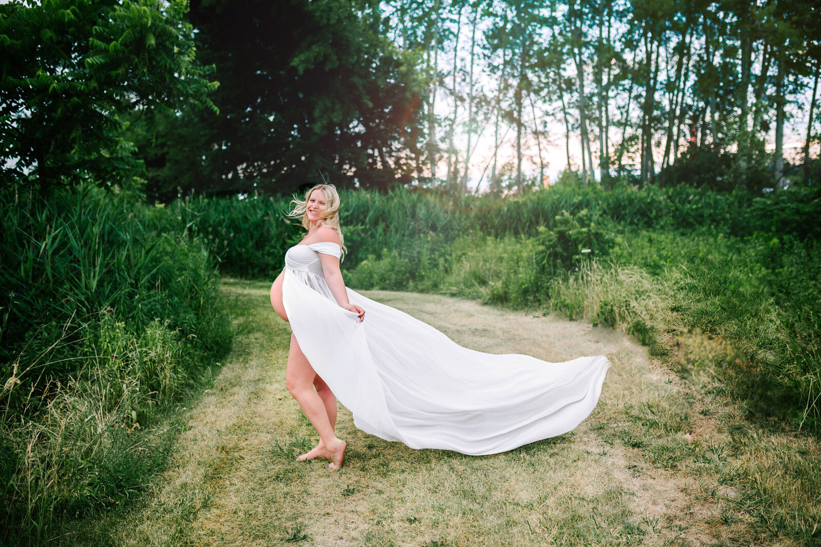 Pregnant mother in long flowing white dress stands with wind blown hair in a grass field