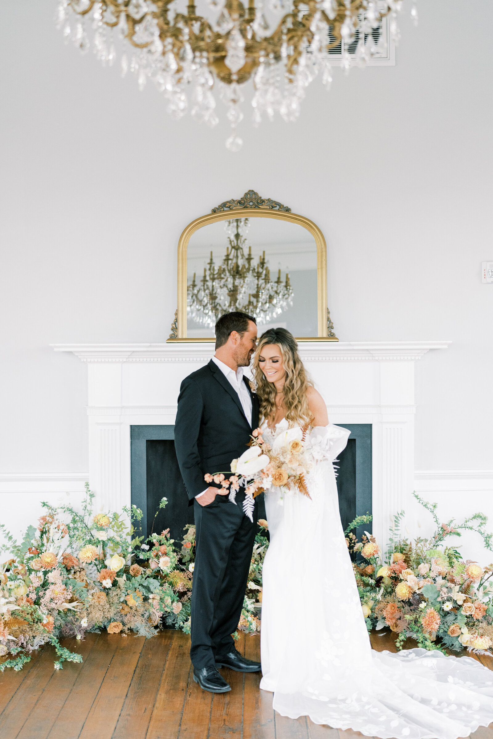 bride and groom standing in front of a fireplace with florals all around them laughing