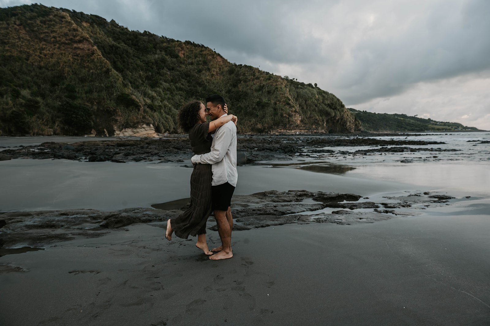 A couple embraces on a rocky beach along the green cliffs, with cloudy skies above—capturing the spirit of an Oregon coast photography retreat for wild-hearted artists.