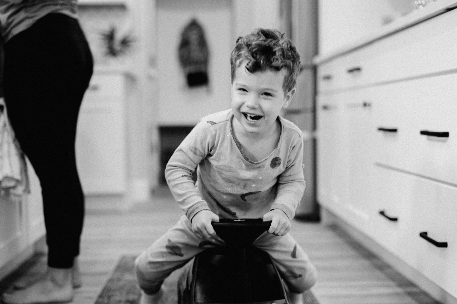 Little boy laughs as he rides a toy car through the kitchen