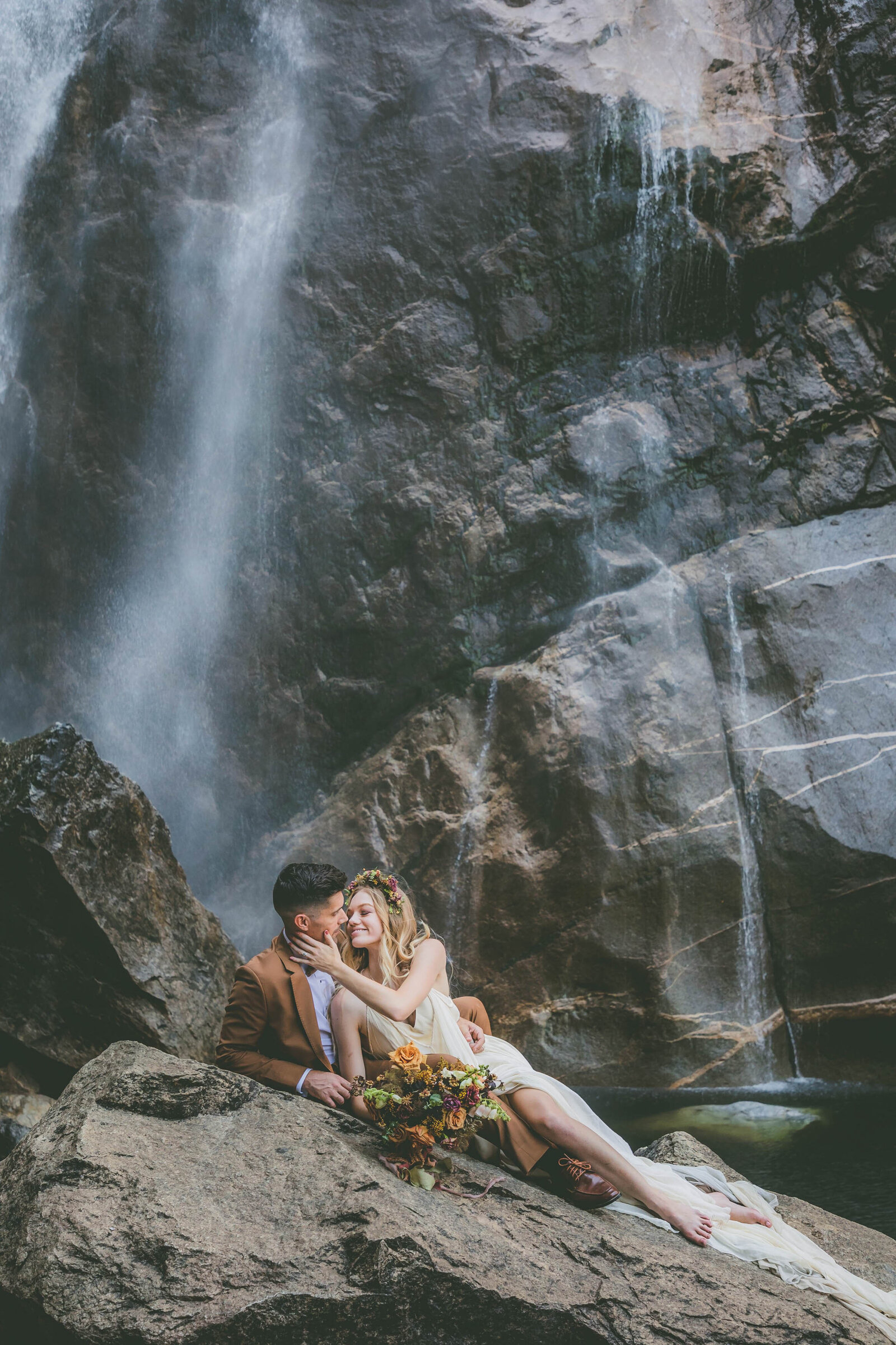 A bride holds her grooms chin as they lay down near a Yosemite waterfall.