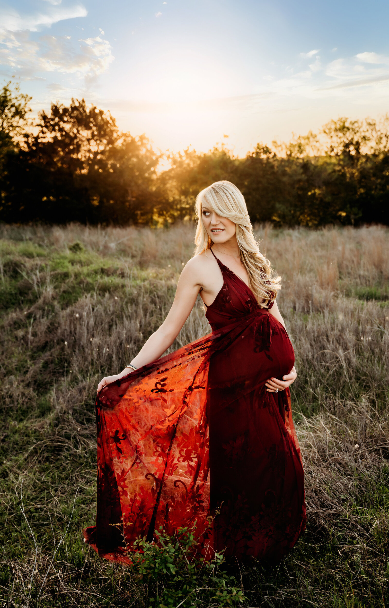Maternity Photographer, a woman in dress, holds its tail in a field, she is pregnant
