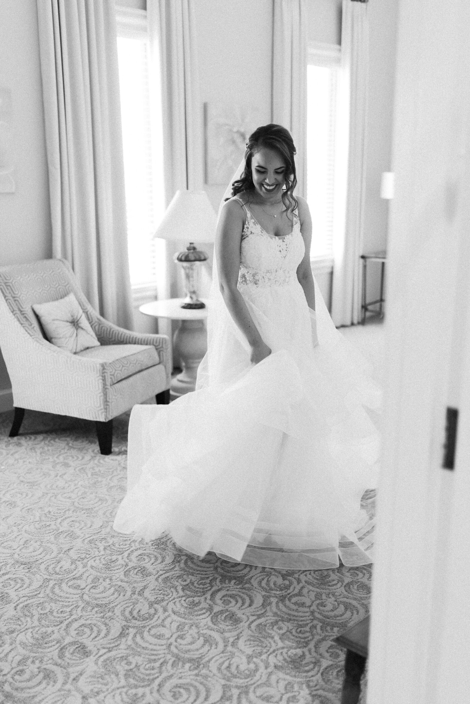 black and white photograph of bride, twirling the bottom of her dress in the dressing room. She is laughing and having fun.