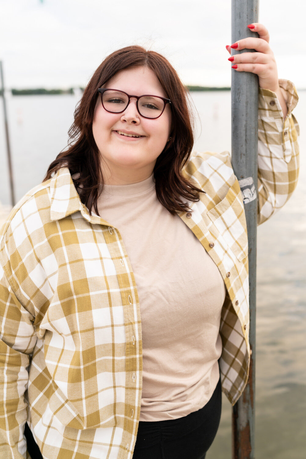 Plus size senior girl smiles with glasses on in front of a lake in Wayzata, Minnesota.