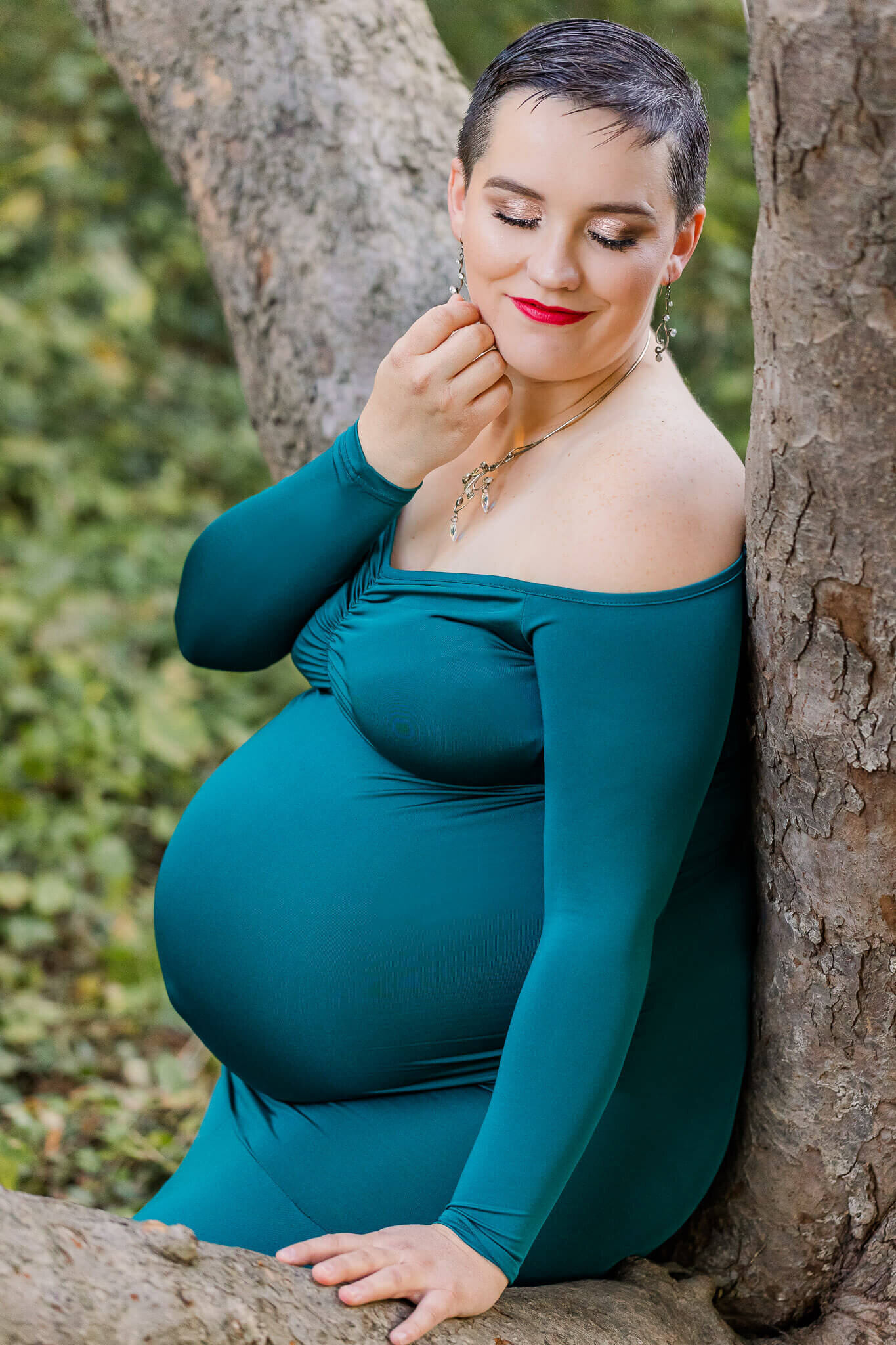 A woman sitting in a tree wearing a teal dress during her maternity photos at an Alexandria park.