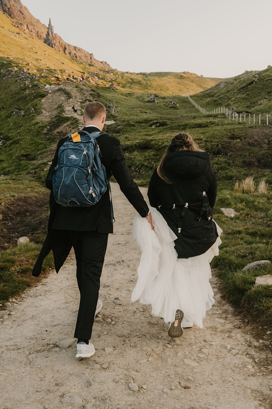 A couple in formal wear and backpacks walk up a rocky, grassy path toward a hill, holding hands—capturing the spirit of a Switzerland photo retreat in the Alps for adventurous photographers.