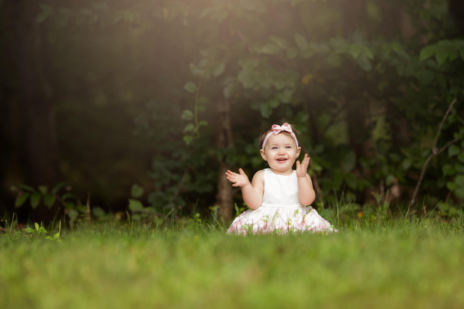 One year old girl sitting in grasss smiling with hands up ready to clap