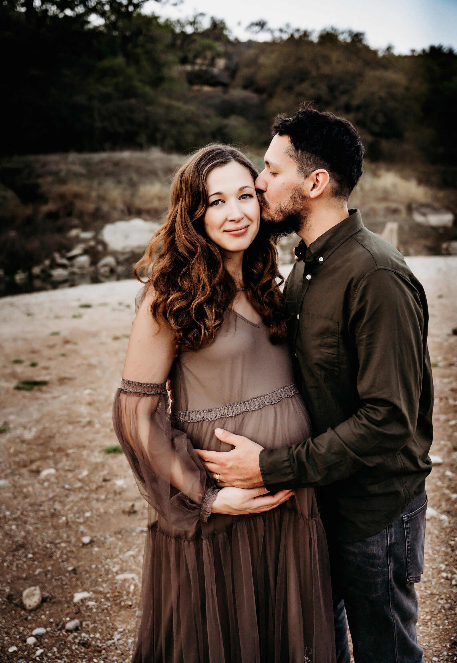 Maternity Photographer, a husband kisses and embraces his pregnant wife at the river