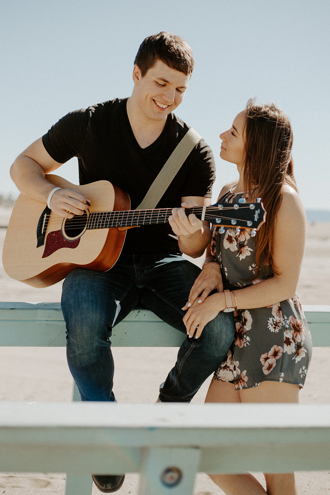 Guy playing an acoustic guitar while his girlfriend smiles at him during their engagement session