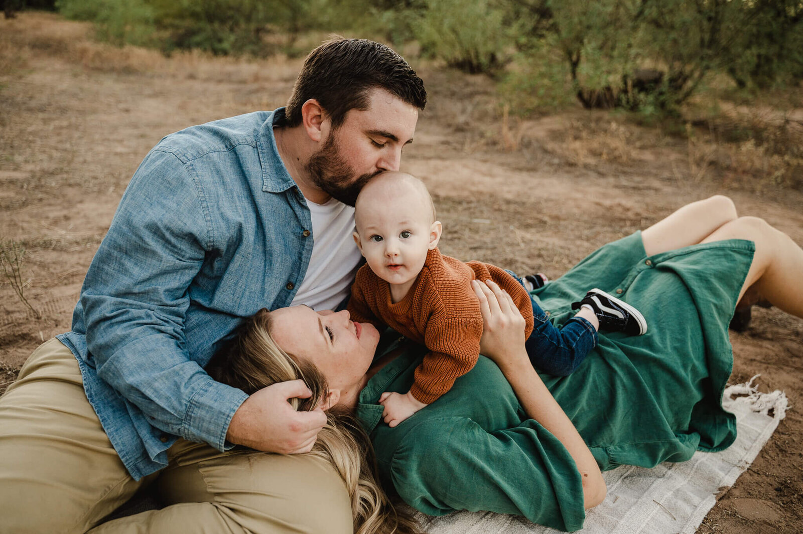mom and dad snuggling on a throw  on a trail with their 1 year old son  amongst desert trees