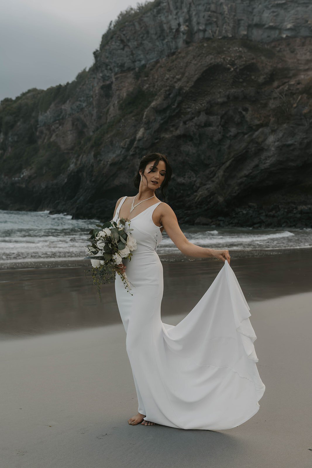 Woman in a white wedding dress holding a bouquet, standing barefoot on a beach with rocky cliffs behind her—an inspiring scene from an Oregon coast photography retreat for wild-hearted artists.