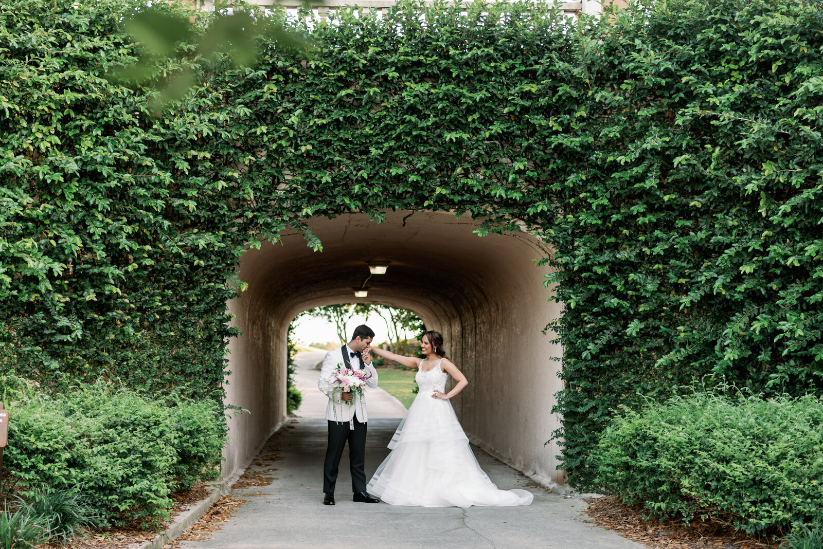 Groom is kissing brides hand while standing in front of an ivy covered tunnel